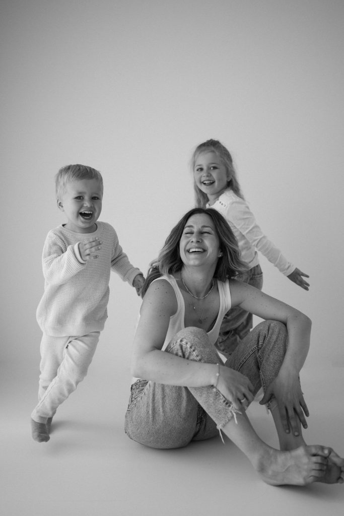 Motherhood Fotografie in Freiburg – Mutter mit zwei Kindern im monochromen Studio, voller Lebendigkeit und Freude